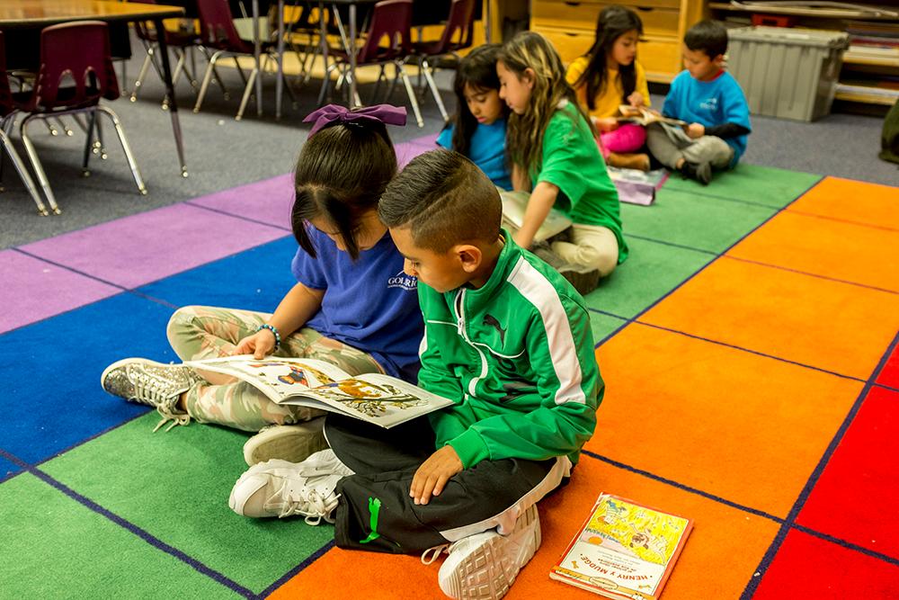 First graders sit cross legged on a multi-colored floor reading in groups of two.