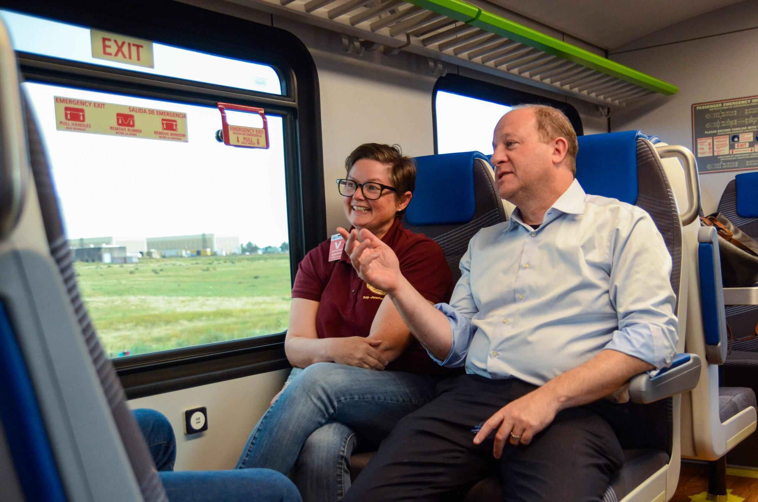 A woman sits next to a man near a window on a train. The man wearing a button down shirt with black pants points out the window. The woman in a red shirt and jeans looks where he's pointing.