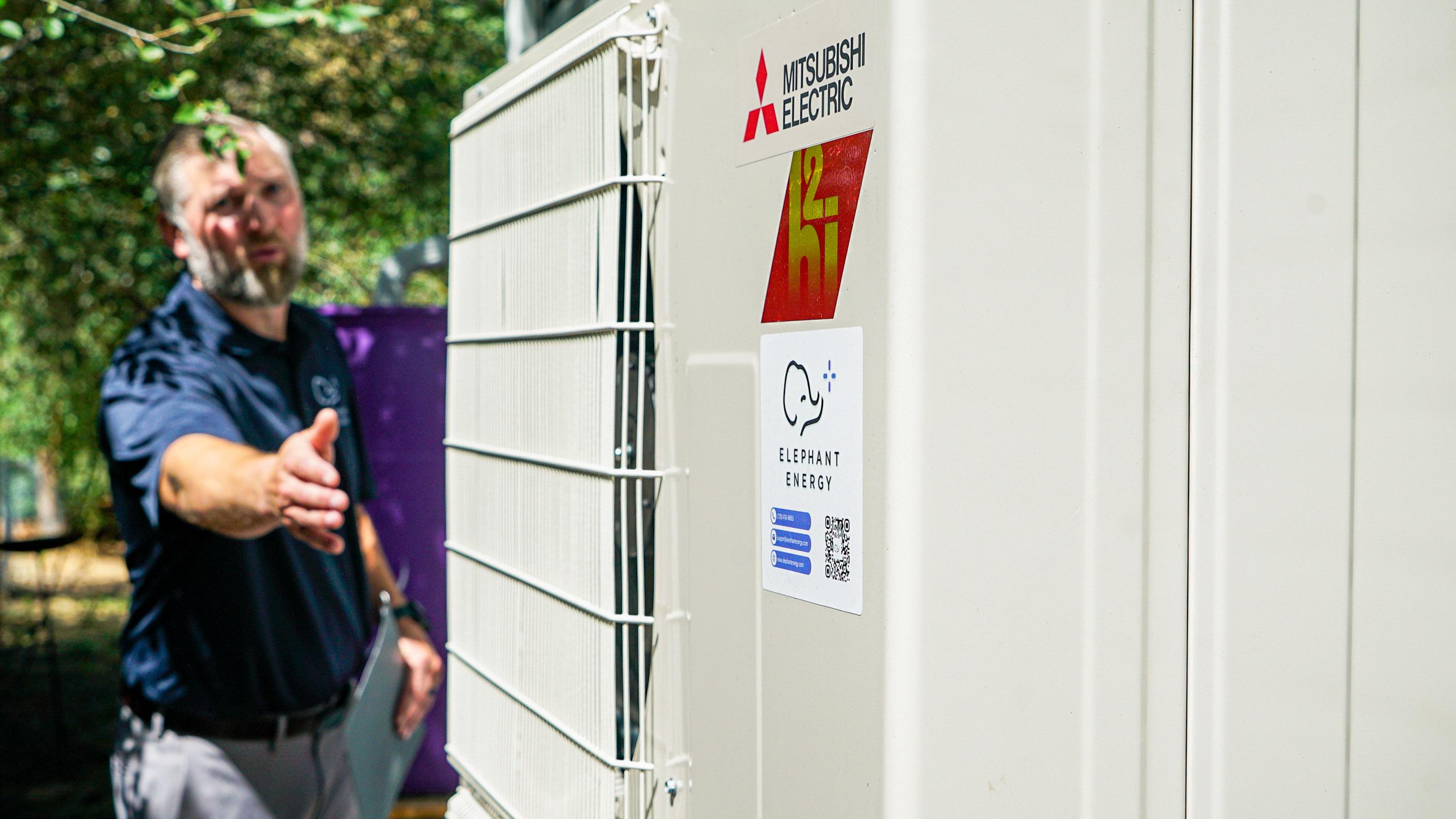 A worker in a blue shirt points at a heat pump installed outside a Denver home.