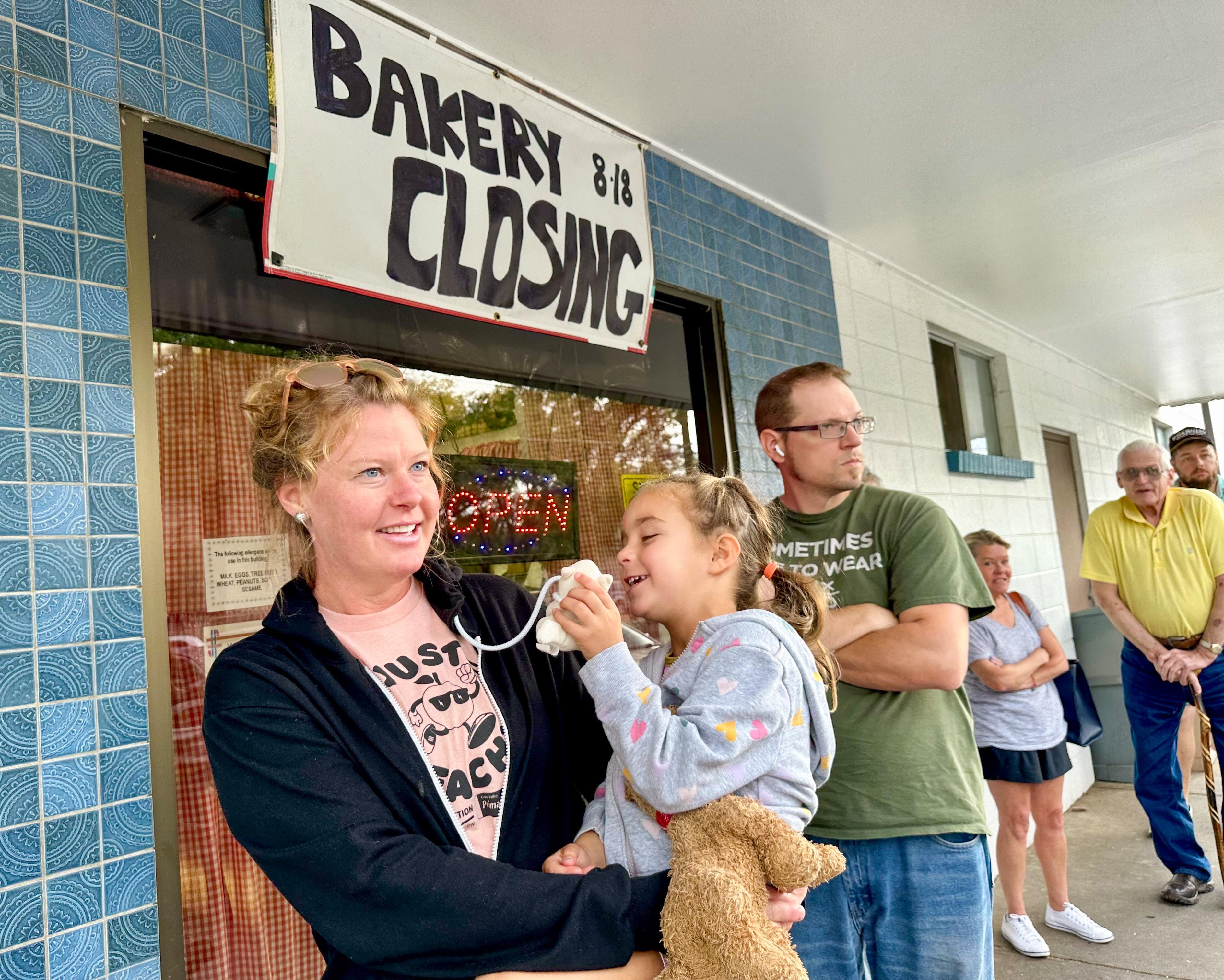 A woman holds her daughter in a bakery line underneath a sign announcing its closing.