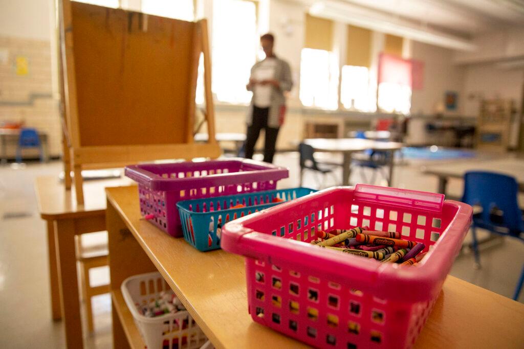 In the foreground, three baskets on top of a brown bookshelf. In the background blurred slightly is a classroom with desks, chairs, and a woman standing near open windows with light coming in.