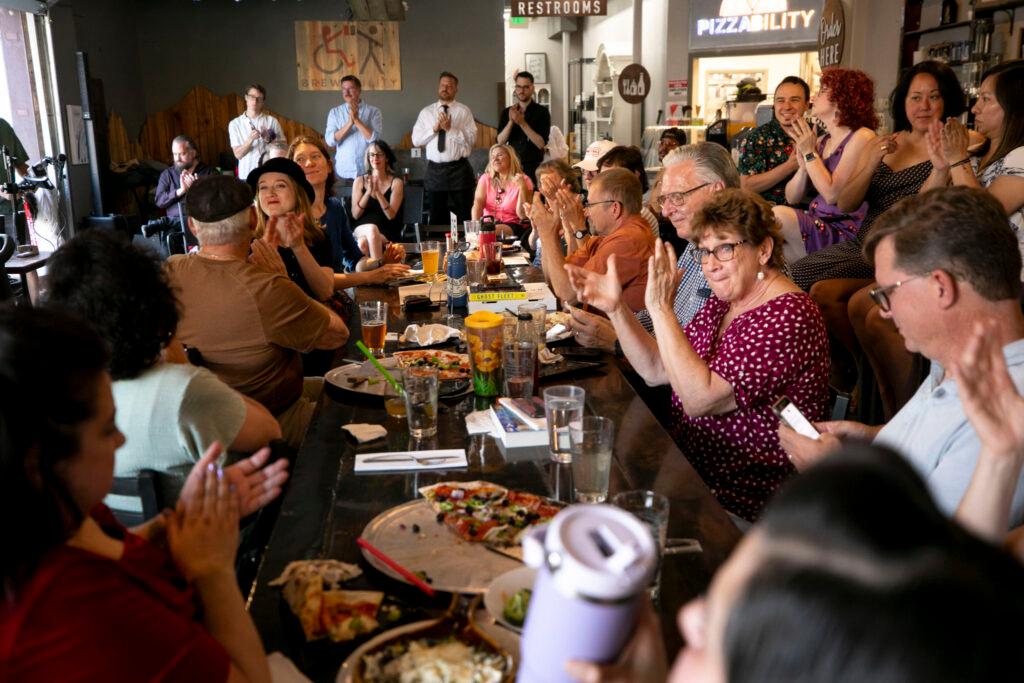 People sitting in a packed restaurant, most around a big table in the middle of the frame, clap and smile.