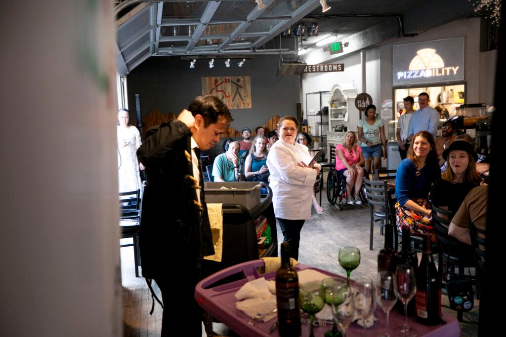 A woman dressed as a chef grimaces at a man dressed as a waiter, as the restaurant full of people around them watch.