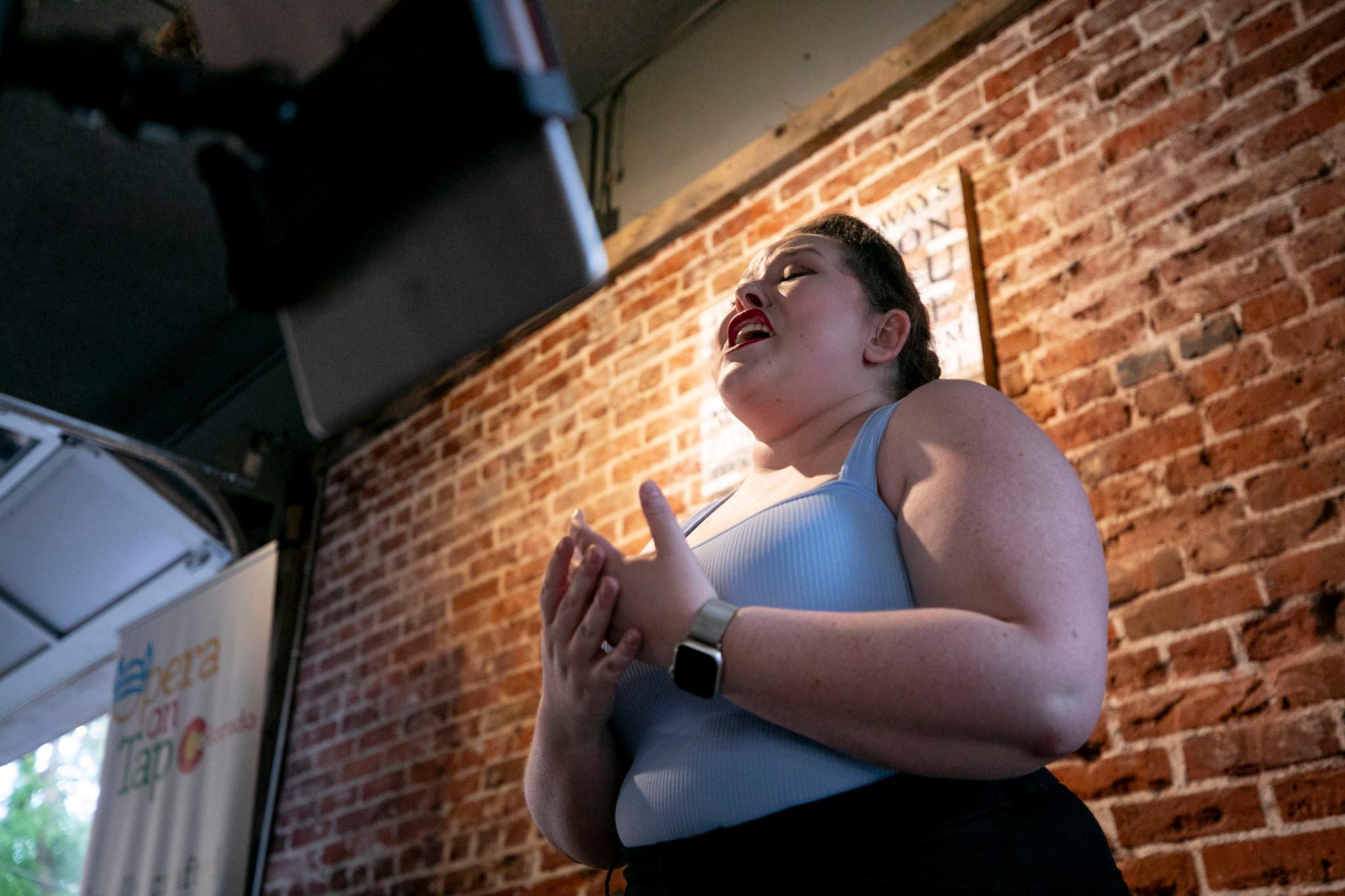 A woman in a tank top sings, her mouth open, her eyes closed and her hands pulling at invisible air.