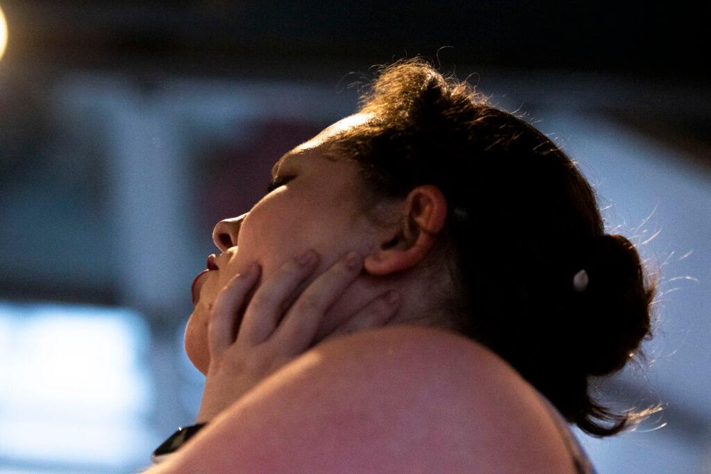 A woman in a tank top sings, her mouth open, her eyes closed and her hands gripping the sides of her jaw.