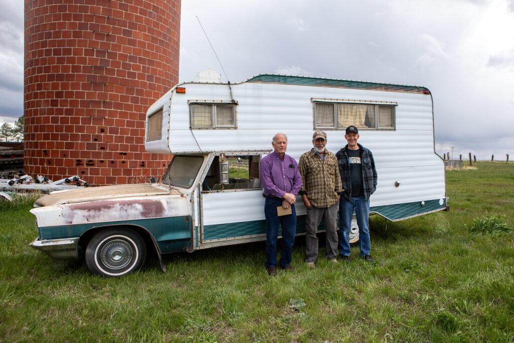 Three men stand in front of a car with a camper build into it.