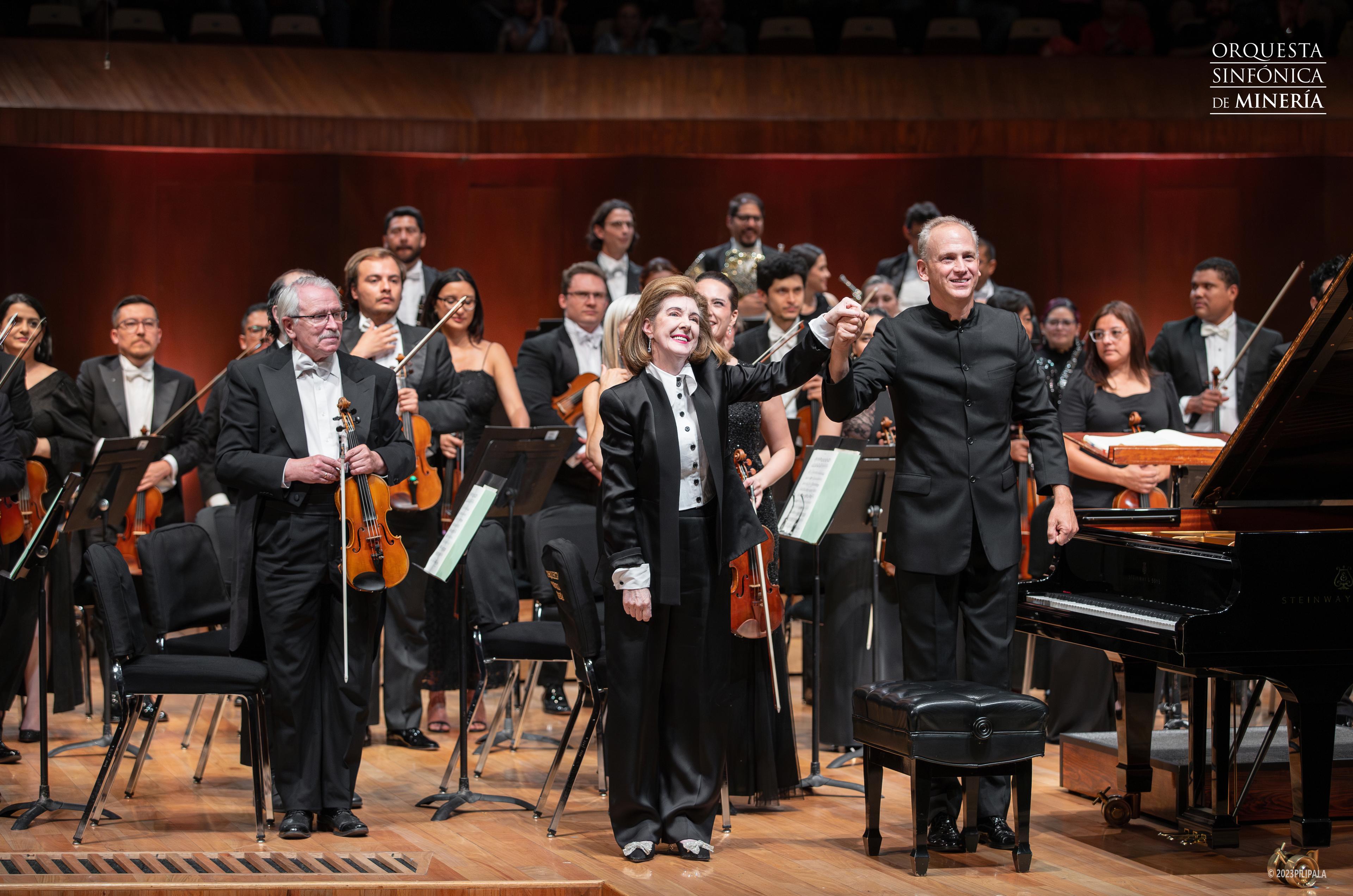 A male conductor and female pianist stand in front of an orchestra with the hands together held high.