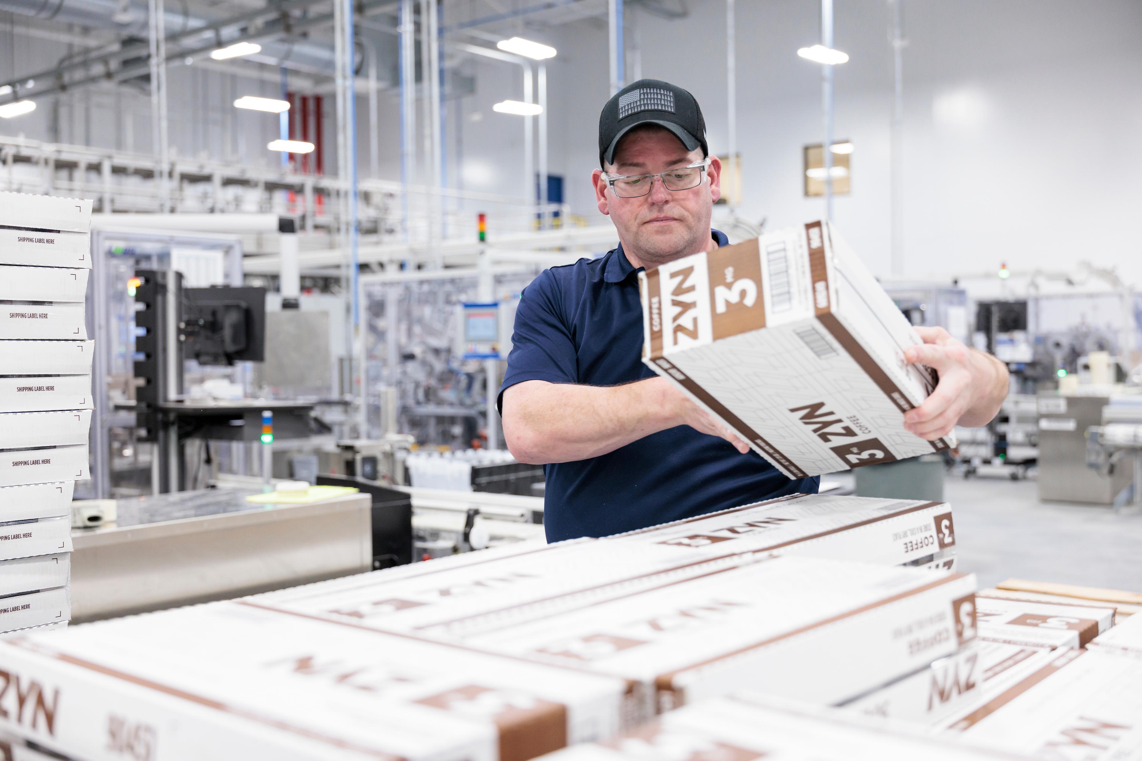 Man in a manufacturing facility wearing a blue shirt and blue hat and goggles prepares to lay down a box on top of other boxes stacked. It's a very sterile looking white background with other boxes and machinery.