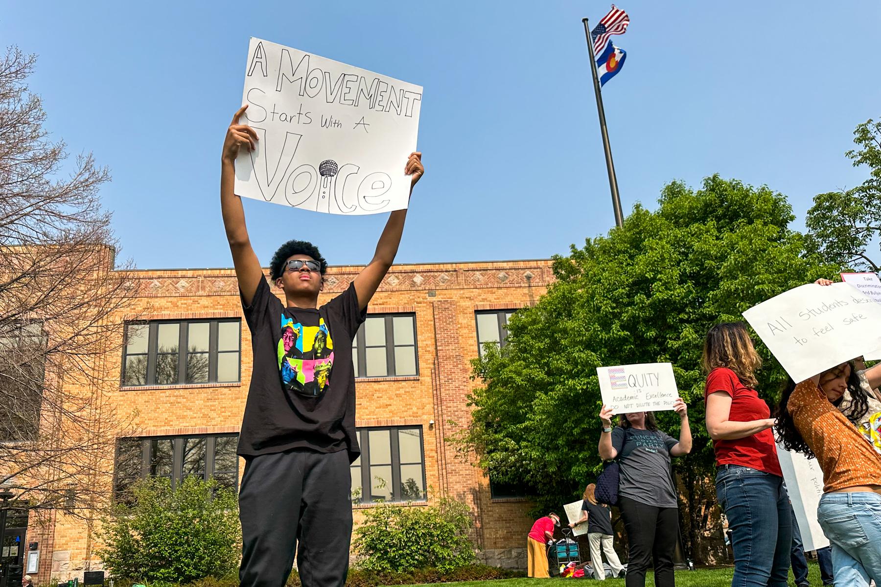 A student in a black t-shirt holds a sign up that reads "A Movement Starts with a Voice" during a protest.