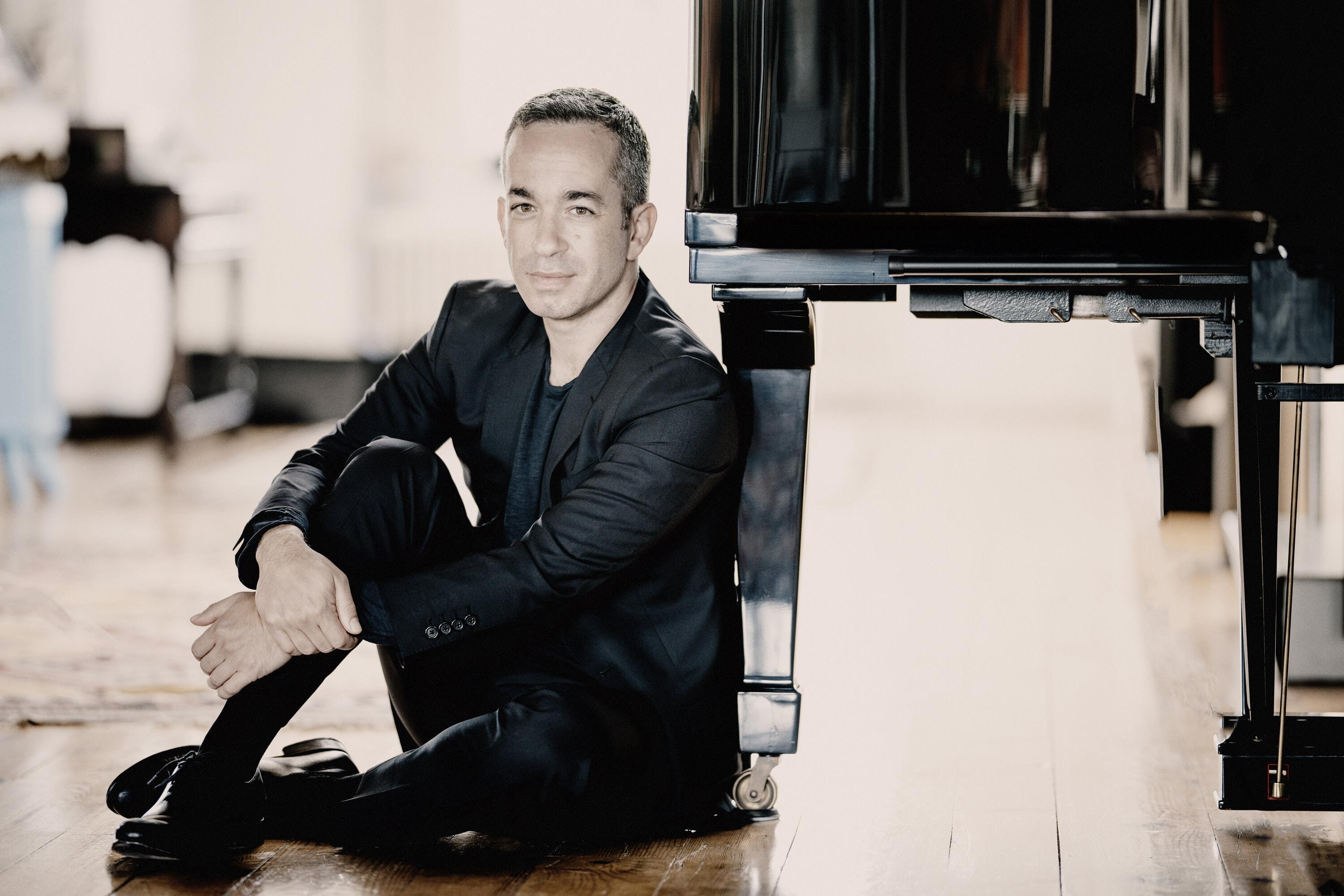 Pianist Inon Barnatan sits on the ground next to a grand piano, looking straight into the camera.